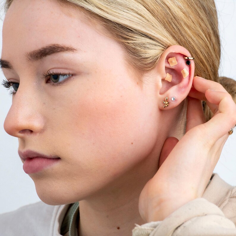 May include: A close-up of a person's ear with multiple gold earrings. The earrings include a small star, a small circle, and a larger hoop with a small black bead. There are also two small square-shaped adhesive bandages on the earlobe.