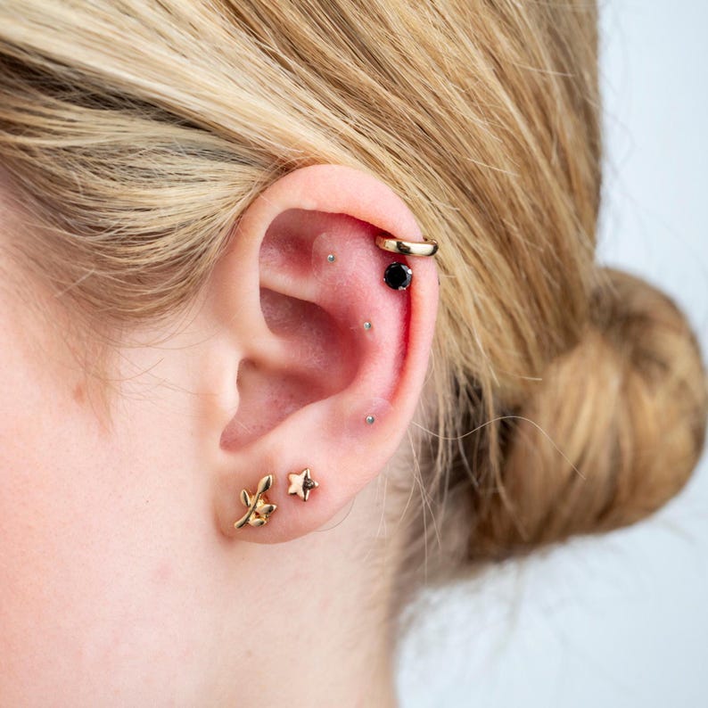 May include: A close-up of a person's ear with multiple piercings. The ear is pierced with a small gold star, a small gold leaf, a small black gem, and a gold hoop.