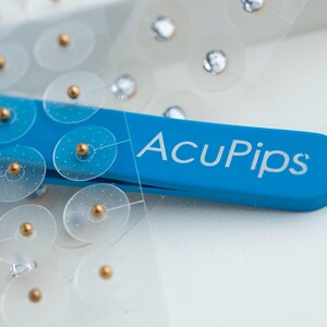 May include: A close-up of a blue plastic tool with the text "AcuPip's" printed on it. The tool is holding a sheet of clear plastic with small, round, adhesive patches with gold dots in the center.
