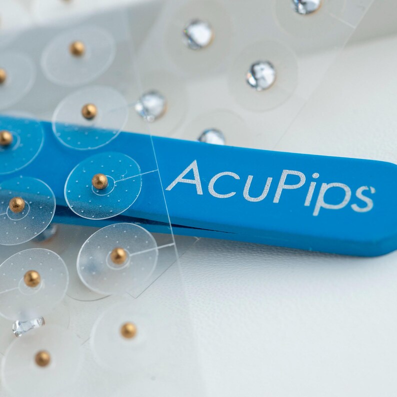 May include: A close-up of a blue plastic tool with the text "AcuPip's" printed on it. The tool is holding a sheet of clear plastic with small, round, adhesive patches with gold dots in the center.