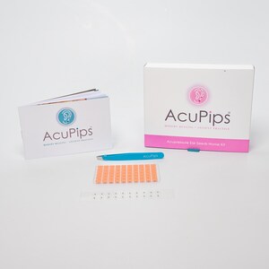 May include: A white box with pink accents and the text "AcuPips" and "Acupressure Ear Seeds Home Kit".  A blue tweezers with the text "AcuPips" is next to the box.  A booklet with the text "AcuPips" is on the left.  A sheet of orange ear seeds is on the table.