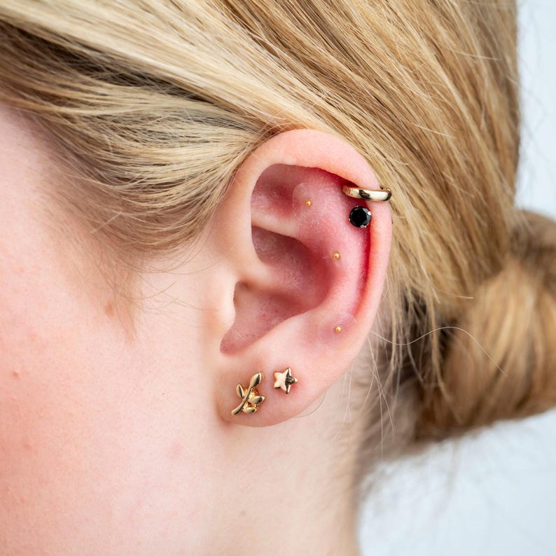 May include: A person's ear with multiple gold earrings. The earrings include a small leaf design, a star, and a hoop with a black gemstone.