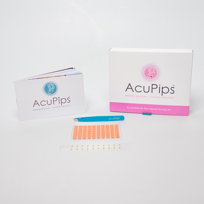 May include: A pink box with the text "AcuPips Modern Healing - Ancient Practice" and "Acupressure for Seeds Home Kit".  A blue tweezers with the text "AcuPips" is next to the box.  A booklet with the text "AcuPips Modern Healing - Ancient Practice" is on the left.  There are two sets of ear seeds on a white background.