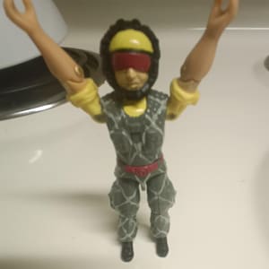 May include: A vintage action figure of a soldier in a gray and green camouflage uniform with a red belt. The figure has a yellow helmet with a red visor and is posed with arms raised.