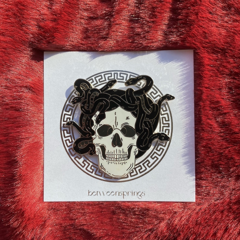 May include: A gold-colored enamel pin featuring a white skull with black snakes for hair. The skull is surrounded by a gold-colored Greek key pattern. The text "betweensprings" is printed below the pin.