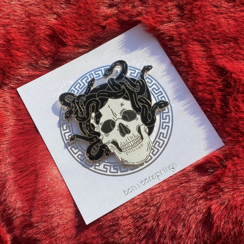 May include: A black and white enamel pin featuring a skull with snakes wrapped around it. The pin is set against a silver background with a Greek key border. The text "bonvsprings" is printed in the bottom right corner.