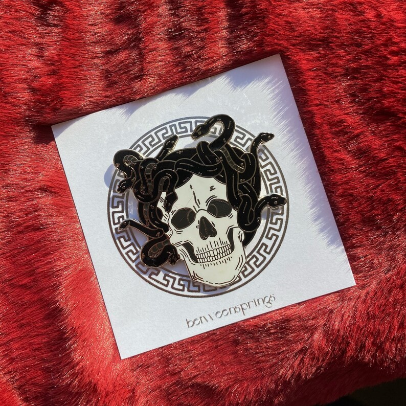 May include: A black and white enamel pin featuring a skull with snakes for hair. The skull is surrounded by a gold-colored Greek key pattern. The pin says "betweensprings" at the bottom.
