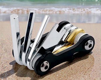 Könnte beinhalten: Ein kompaktes Multitool mit silbernen und goldenen Elementen, das auf einem Sandstrand liegt. Das Werkzeug mit schwarzem Gehäuse ist für die Fahrradwartung konzipiert. Im Hintergrund sind Meer und Himmel zu sehen.