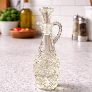 Peut inclure: Une burette en verre transparent avec un motif floral décoratif. La burette a une poignée et un bouchon. L'arrière-plan comprend une cuisine avec d'autres ustensiles et ingrédients.