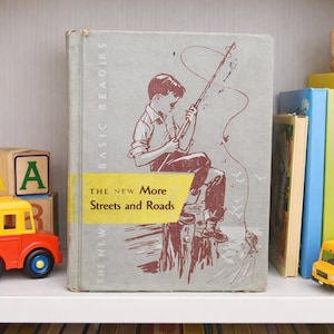 May include: A shelf displays a vintage children's book titled "The New More Streets and Roads" with an illustration of a boy fishing. Other items include toy buses, alphabet blocks, and a teddy bear, suggesting a children's theme.