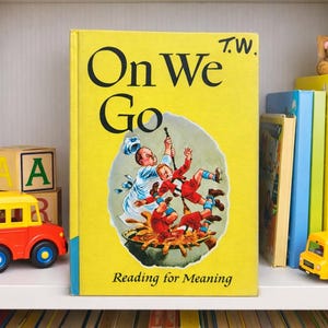 May include: A yellow children's book titled "On We Go" with an illustration of people falling into a pot. The book is on a shelf with other books, toy blocks, a teddy bear, and toy vehicles.