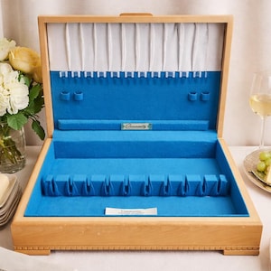 May include: Wooden cutlery chest with a bright blue felt interior. The chest has multiple compartments for storing utensils. The interior is lined with a light blue felt. The chest is open, revealing the interior.