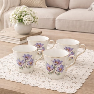 May include: Four white teacups with gold rims and handles, each decorated with a purple and green floral design. The cups are arranged on a white lace doily on a wooden table. A vase of white flowers sits in the background.