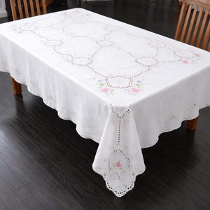 May include: A white rectangular tablecloth with floral embroidery and cutwork details. The tablecloth is draped over a wooden table, showcasing a delicate design with pink, blue, and green floral accents.
