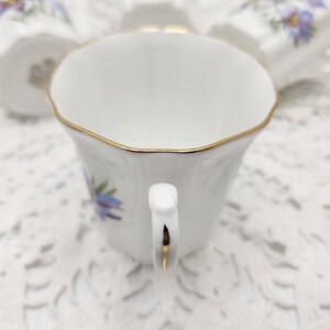 May include: White teacup with a gold rim and handle, adorned with a blue floral design. The cup is part of a tea set, showcasing a delicate and elegant aesthetic. The handle and rim are accented with gold trim.