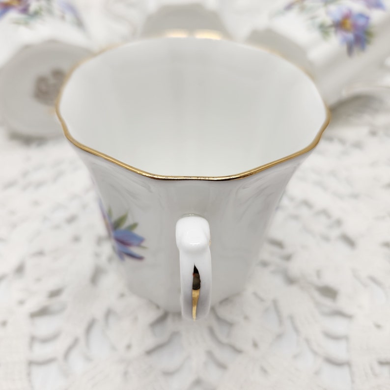 May include: White teacup with a gold rim and handle, adorned with a blue floral design. The cup is part of a tea set, showcasing a delicate and elegant aesthetic. The handle and rim are accented with gold trim.