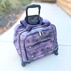 Peut inclure: Valise à roulettes violette avec un motif texturé en forme de peau de crocodile. La valise a une poignée rétractable et le mot "TRACKER" est visible sur les fermetures éclair et la poignée. Le sac a deux roues et une poignée supérieure.