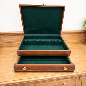 May include: A wooden jewelry box with a dark brown finish and a green velvet interior. The box is open, revealing multiple compartments and a pull-out drawer. Gold-colored hardware accents the box.