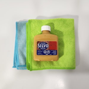 May include: A bottle of Silvo metal polish, a gold colored bottle with a red and white label, sitting on top of two microfiber cloths, one blue and one green.