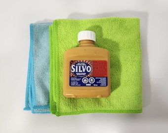 May include: A bottle of Silvo metal polish, a gold colored bottle with a red and white label, sitting on top of two microfiber cloths, one blue and one green.