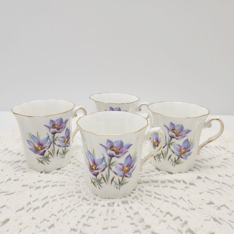 May include: Four white porcelain teacups with gold trim and handles. Each cup features a floral design with purple and blue flowers. The cups are arranged on a white lace tablecloth.