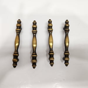 May include: Four vertical cabinet pulls with an antique brass finish. Each handle features a central elongated body with rounded, tiered decorative accents at the top and bottom ends, creating a uniform, symmetrical design for furniture hardware.