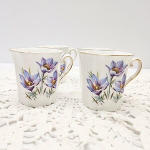 May include: Four white porcelain mugs with a floral design. Each mug features a gold rim and handle, and is decorated with purple and blue flowers with green stems. The mugs are arranged on a white lace surface.