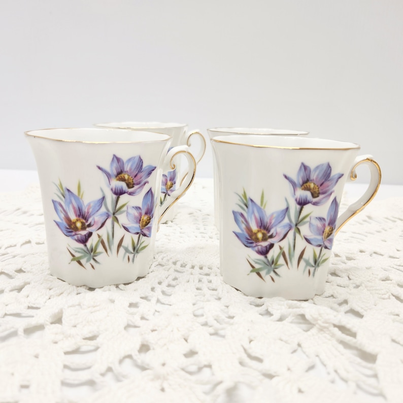 May include: Four white porcelain mugs with a floral design. Each mug features a gold rim and handle, and is decorated with purple and blue flowers with green stems. The mugs are arranged on a white lace surface.