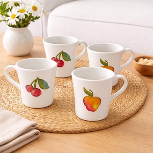 May include: Four white ceramic mugs with fruit illustrations. Two mugs feature red cherries with green leaves, and two have orange and red apples with green leaves. The mugs are on a woven placemat, with a vase of white flowers in the background.