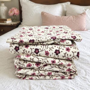 May include: A stack of folded fabric featuring a cream-colored background with a repeating pattern of dark purple and muted pink flowers connected by brown vine-like stems. The textile has a smooth texture and is neatly layered in a vertical pile.