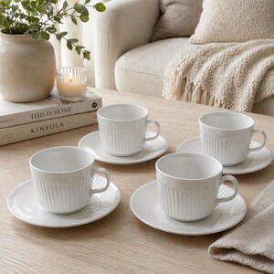 May include: Four white ceramic teacups and saucers arranged on a light wood table. The cups and saucers have a ribbed design. A vase with greenery, a candle, and books are in the background.