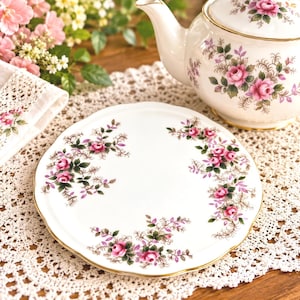 May include: A white ceramic plate with a scalloped edge and a gold-trimmed rim, featuring a delicate floral pattern of pink roses and green leaves arranged in clusters around the center, matching the design on the adjacent teapot.
