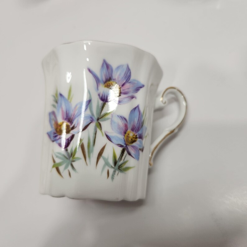 May include: White porcelain teacup with a floral design. The cup features a delicate handle and is adorned with blue and purple flowers with yellow centers and green leaves. The rim and handle are trimmed with gold.