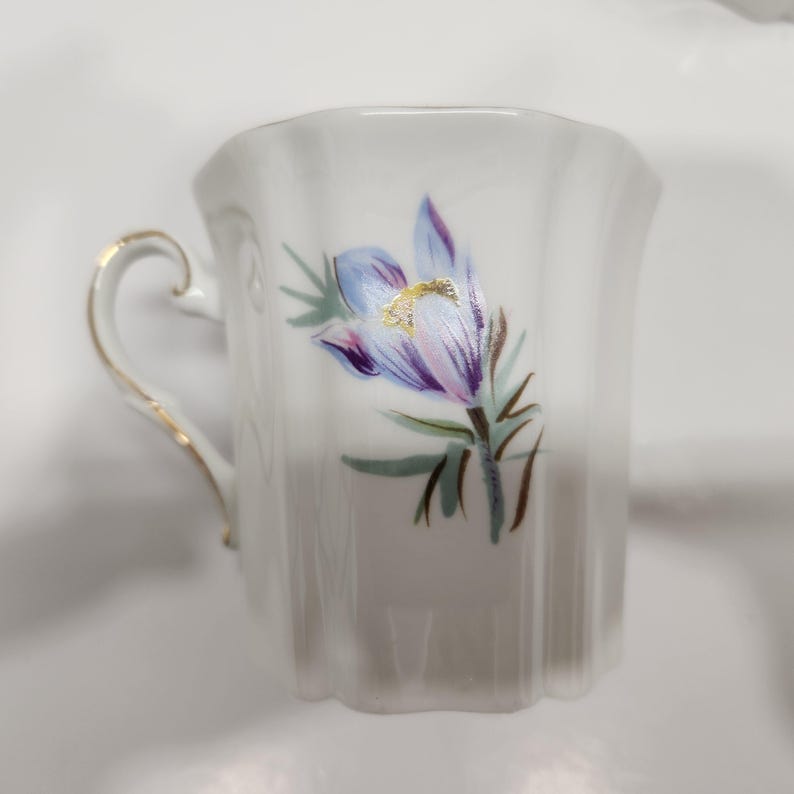 May include: White ceramic teacup with a gold handle and rim. The cup features a hand-painted floral design with blue, purple, and gold accents. The cup has a scalloped edge and a curved handle.