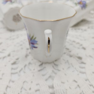 May include: White porcelain teacup with a gold rim and handle, adorned with blue floral designs. The cup is set on a white lace surface, with a matching saucer visible in the background. The teacup has a delicate, vintage aesthetic.