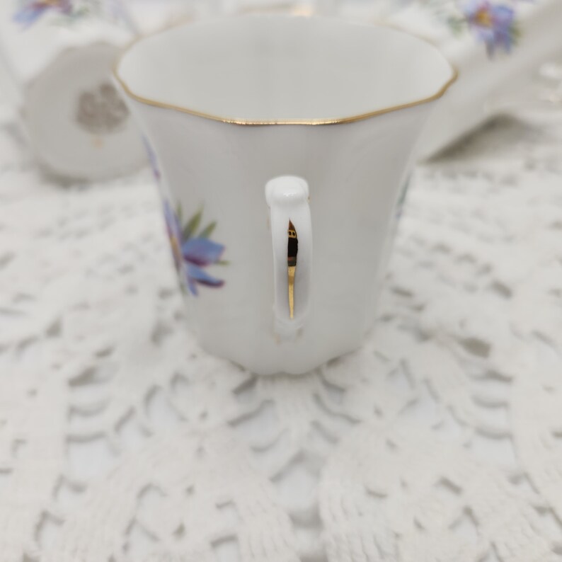 May include: White porcelain teacup with a gold rim and handle, adorned with blue floral designs. The cup is set on a white lace surface, with a matching saucer visible in the background. The teacup has a delicate, vintage aesthetic.