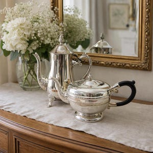 May include: A polished silver tea set, comprising a teapot and a sugar bowl, with intricate designs. The teapot has a curved spout and a decorative handle. The sugar bowl has a black handle. A vase of white flowers and a gold-framed mirror are in the background.