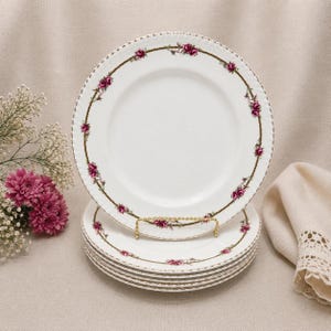 May include: A set of white porcelain plates with a floral rose design. Each plate has a gold rim and a border of pink roses and green leaves. The plates are displayed on a gold plate stand.
