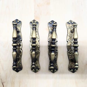 Peut inclure: Quatre poignées de porte en laiton antique avec des détails ornés. Chaque poignée présente un design vertical avec une texture centrale en forme de corde et des embouts décoratifs. Montées sur une surface en bois clair.