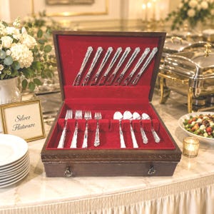 May include: A wooden flatware storage chest with a red velvet interior containing a set of silver-toned utensils. The open lid displays knives, while the base holds forks and spoons in individual slots. The exterior features a dark wood finish.