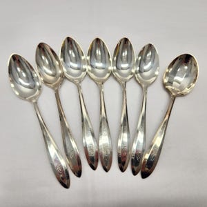 May include: A collection of seven silver spoons displayed on a white background. The spoons feature a traditional design with slender handles and oval bowls. Each spoon has a decorative marking on the handle.