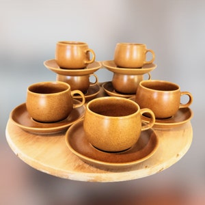 May include: Set of six brown ceramic coffee cups and saucers. The cups and saucers are arranged in a circle on a wooden tray.