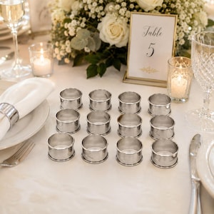 May include: A set of twelve polished silver-toned metal napkin rings arranged in a grid on a white tablecloth. Each ring features a smooth, circular band design. The napkin rings are positioned near a white plate, a glass, and a table sign.