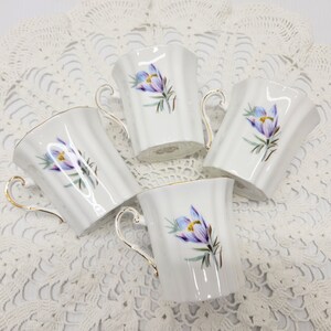 May include: Four white porcelain teacups with a floral design featuring purple and yellow flowers. Each cup has a gold handle and trim. The cups are arranged on a white lace tablecloth.