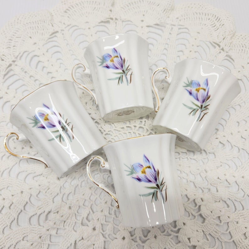 May include: Four white porcelain teacups with a floral design featuring purple and yellow flowers. Each cup has a gold handle and trim. The cups are arranged on a white lace tablecloth.
