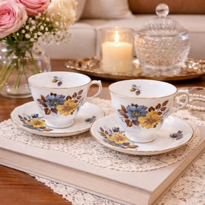 May include: Two white teacups and saucers with blue and yellow floral designs and gold trim. The teacups and saucers are placed on a book and a lace doily. A vase of pink roses and a lit candle are in the background.