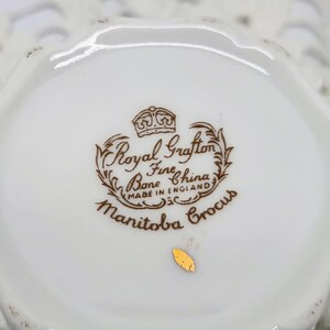 May include: Close-up of a white Royal Grafton Fine Bone China teacup base. The brown text reads "Royal Grafton Fine Bone China Made in England Manitoba Crocus". A small gold sticker is visible.