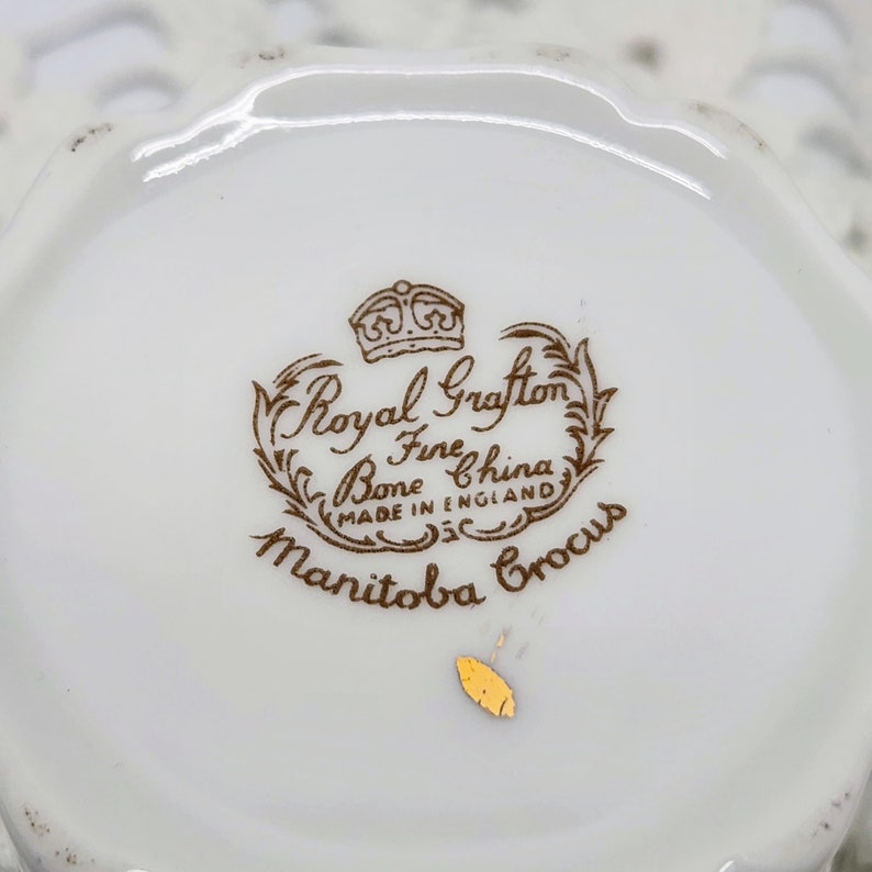 May include: Close-up of a white Royal Grafton Fine Bone China teacup base. The brown text reads "Royal Grafton Fine Bone China Made in England Manitoba Crocus". A small gold sticker is visible.