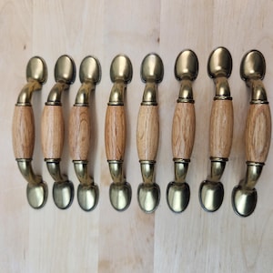 May include: Eight cabinet handles with a brass finish and light wood centres. The handles are curved with oval-shaped ends and a cylindrical wooden centre. The handles are arranged in a row on a light wood surface.
