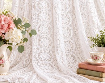 May include: A white lace curtain with a repeating floral pattern hangs as a backdrop. To the left, a ceramic pitcher holds pink and white hydrangeas. To the right, a floral teacup sits atop a stack of two vintage books.